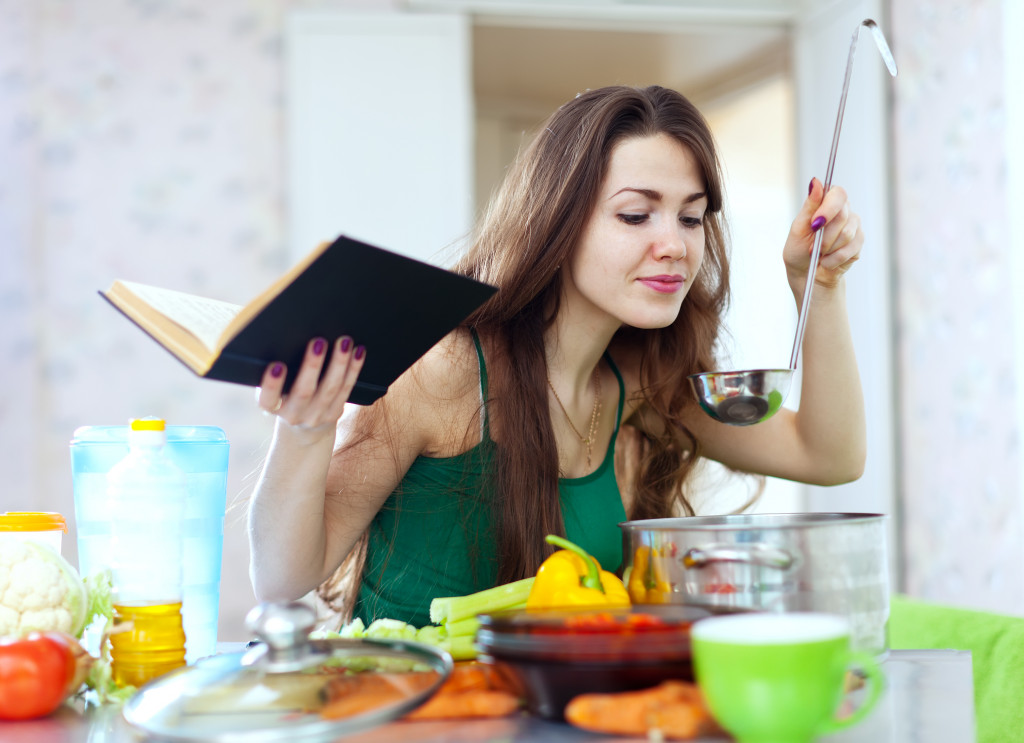 A female home-based restaurant chef is tasting her food with a notepad in her other hand.
