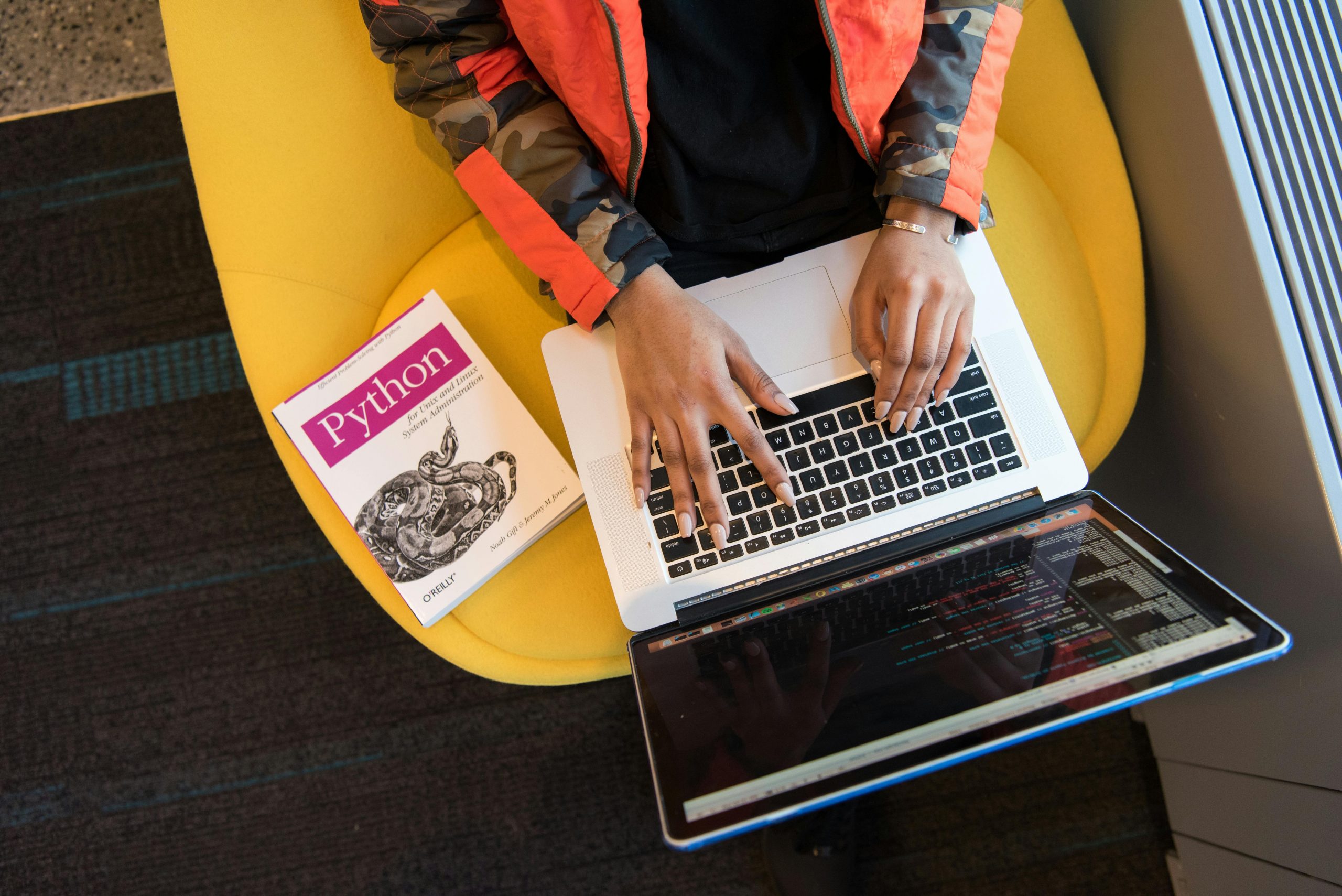 woman typing on a laptop on her lap, with a Python programming book next to her